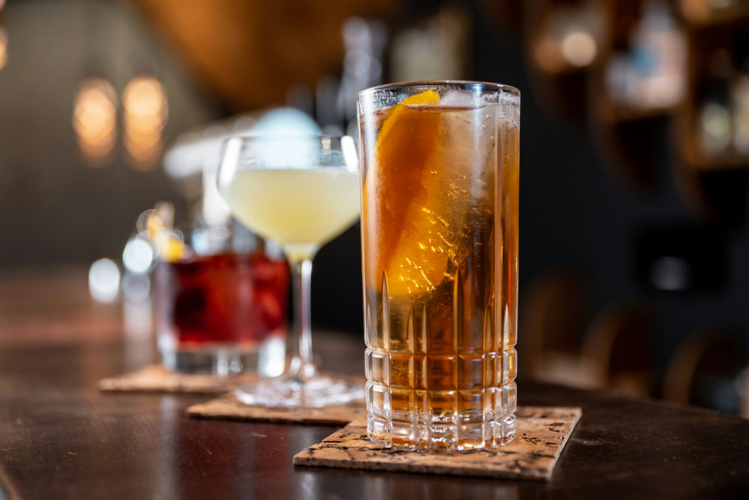 three different classic cocktails sit on a dark wood bar at an event